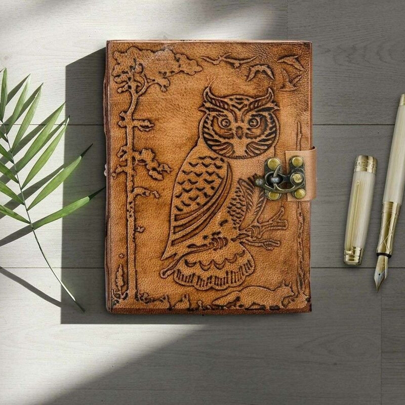 Brown leather notebook with owl design on a wooden surface with pens and a plant.