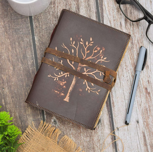 Brown leather notebook with tree design on a wooden surface with a pen and glasses.