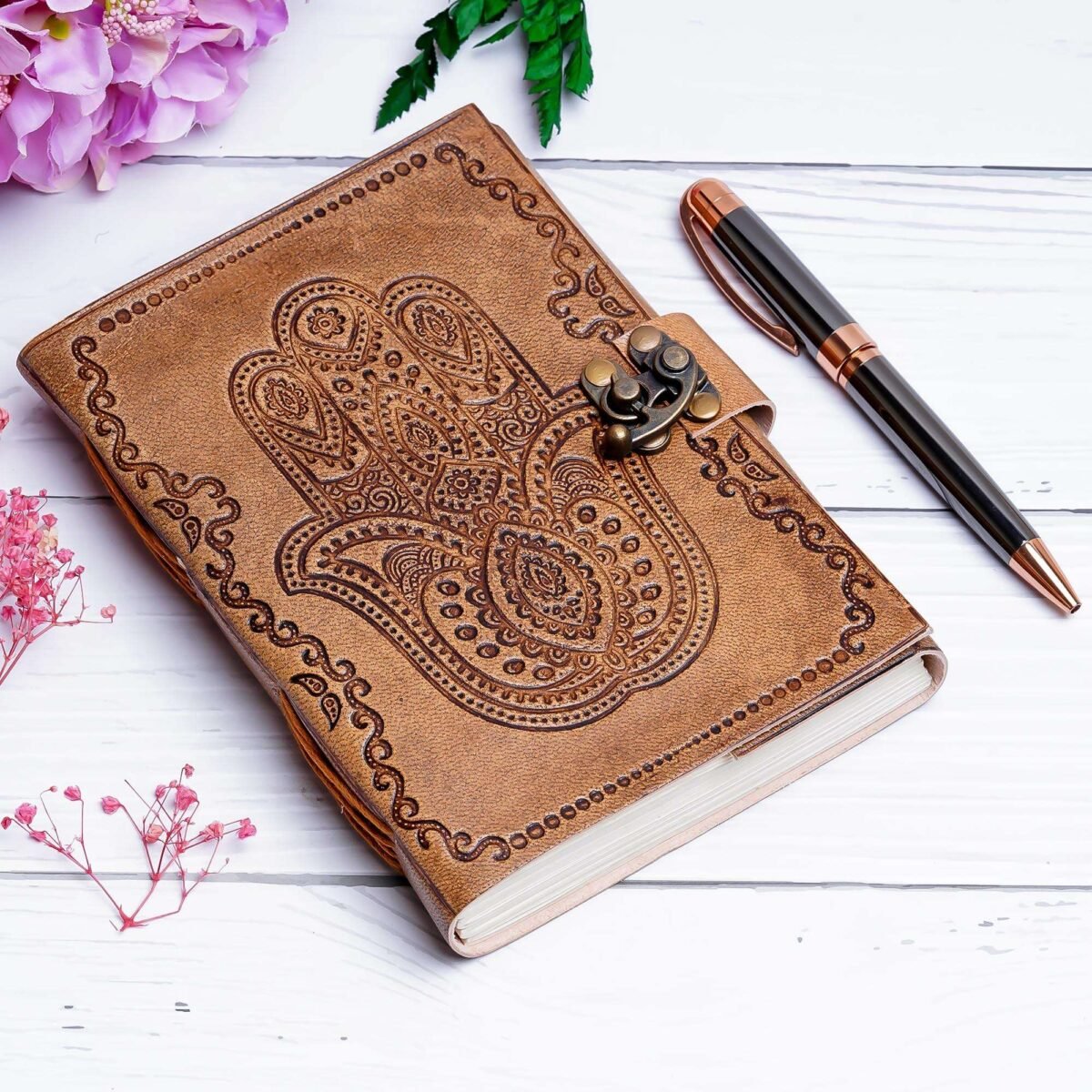 Brown leather notebook with paisley design and pen on a white surface with pink flowers.
